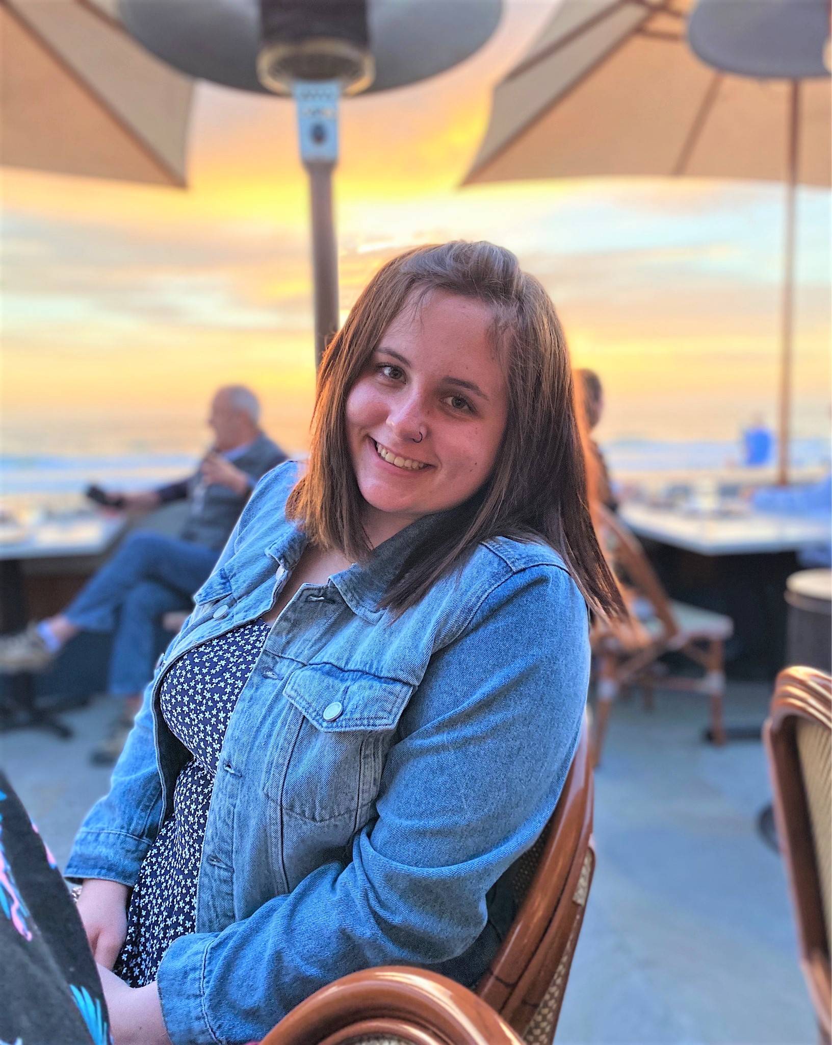Woman with shoulder length brown hair wearing jean jacket by a table with a sunset beach in the background